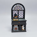A miniature black witch hut figurine with a stained-glass style arched window, a small black witch hat on top, and tiny shelves with tiny accessories, set on a wooden tabletop.