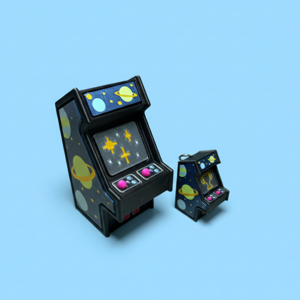 Two miniature arcade machine keychains styled with a space theme, featuring planets, stars, and colorful buttons, displayed on a wooden tabletop.