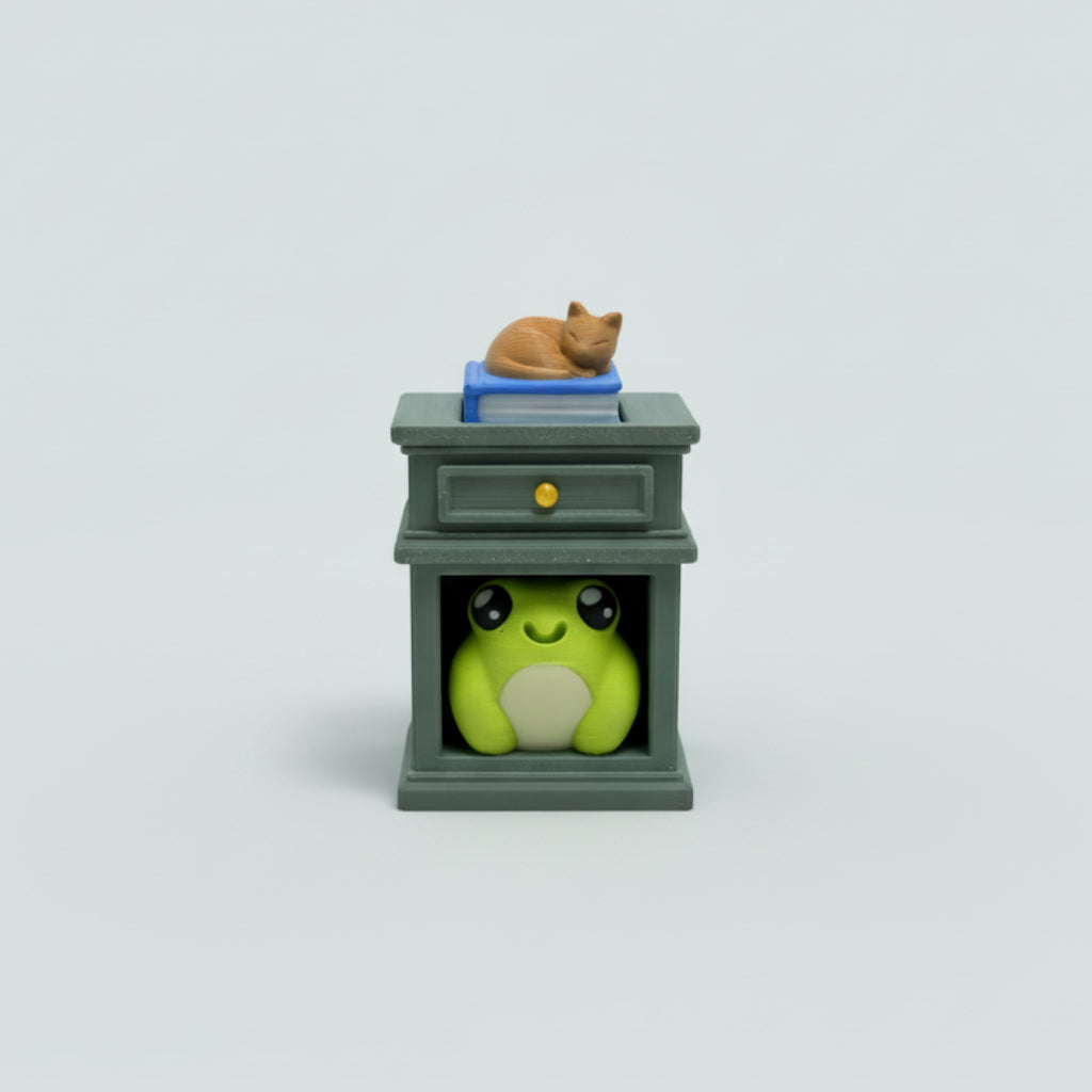 A small decorative cabinet figurine on a wooden tabletop, featuring a green frog peeking out from beneath and a blue book with a sleeping orange cat on top.