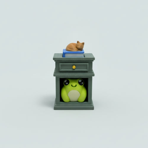 A small decorative cabinet figurine on a wooden tabletop, featuring a green frog peeking out from beneath and a blue book with a sleeping orange cat on top.