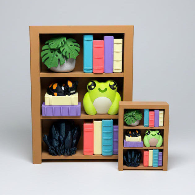 A miniature toy bookshelf set on a wooden surface, featuring a larger brown bookshelf with colorful toy books, a green frog figure, and small plants, alongside a smaller matching bookshelf.
