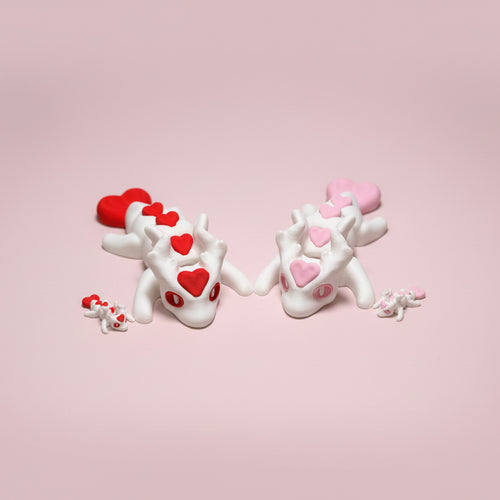 Two 3d printed dragon figures holding hands with heart-shaped eyes on a pink surface
