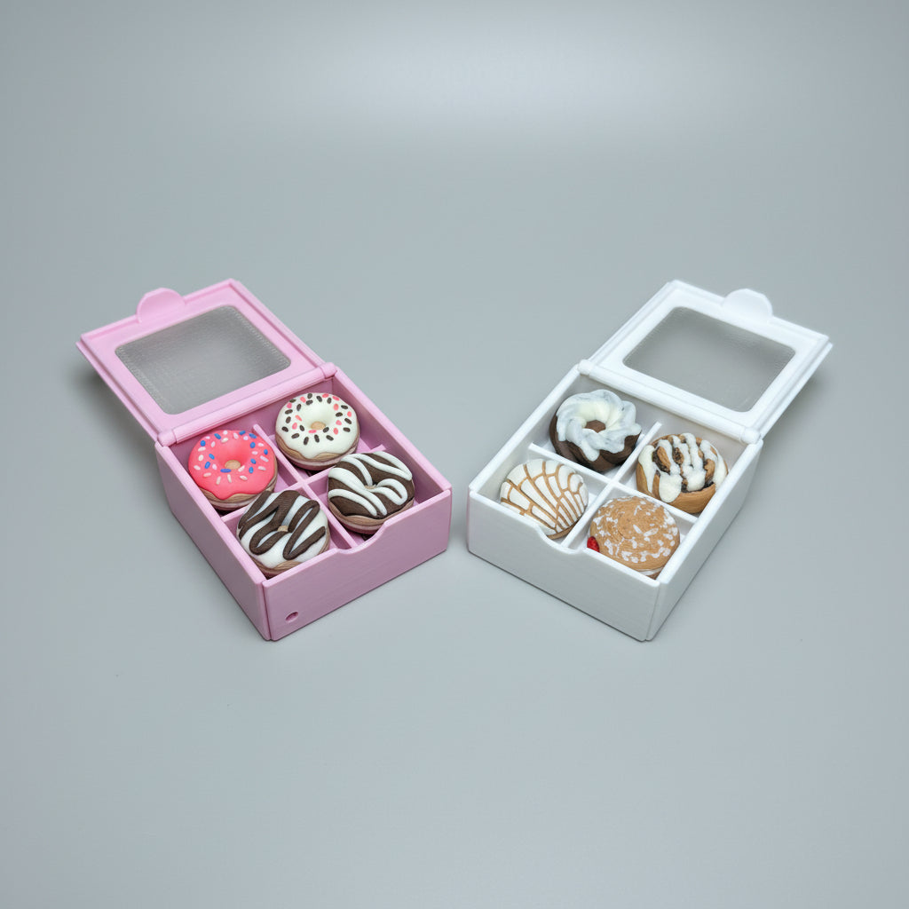 Two miniature donut-shaped boxes on a light wood surface, one pink and one white, each containing six small donut-shaped items with different frosting and topping designs.