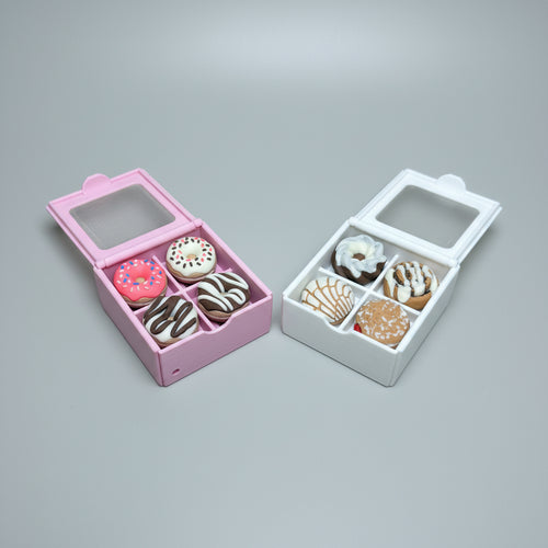 Two miniature donut-shaped boxes on a light wood surface, one pink and one white, each containing six small donut-shaped items with different frosting and topping designs.