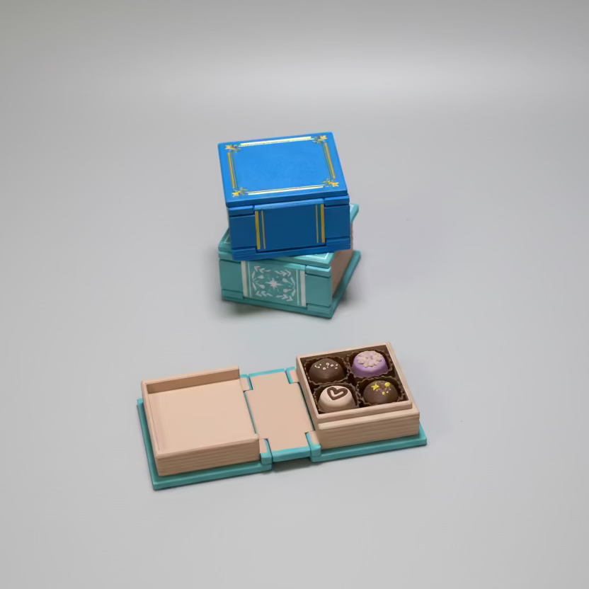 Blue and green book style box with open lid revealing small round pretend chocolate truffles on a grey surface.